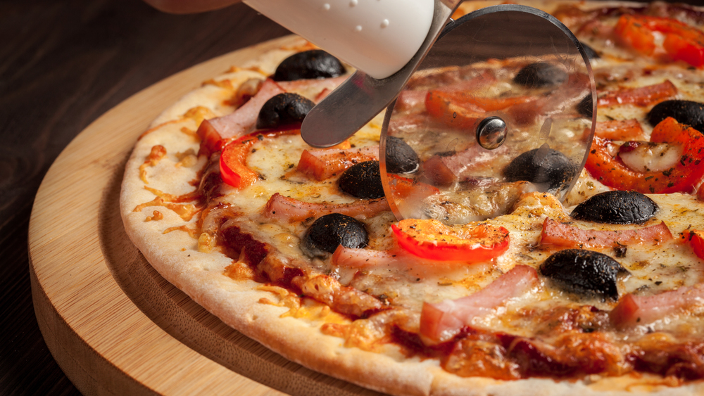 Letterbox panorama of pizza cutter (wheel) slicing ham pizza with capsicum and olives on wooden board on table. Pizza cutter wheel slicing ham pizza