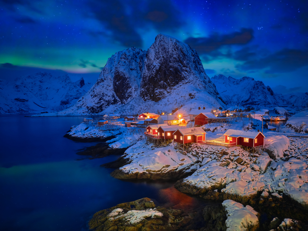 Famous tourist attraction Hamnoy fishing village on Lofoten Islands, Norway with red rorbu houses in winter snow illuminated in the evening with Aurora Borealis. Hamnoy fishing village on Lofoten Islands, Norway