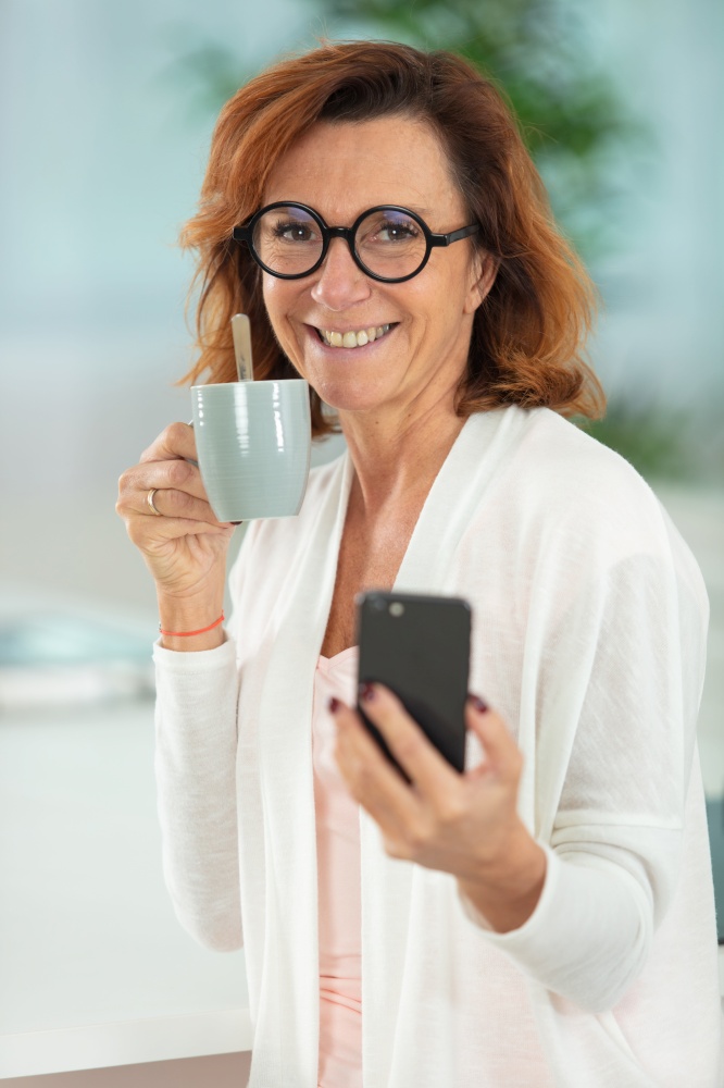 mature woman holds coffee and uses smartphone in the home