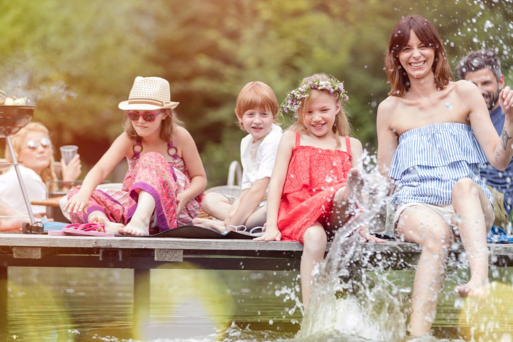 Smiling woman and daughter splashing water in lake while sitting on pier with family during summer picnic