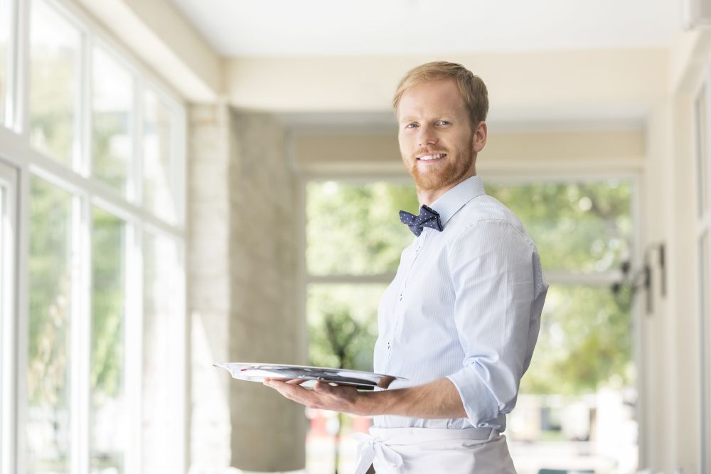 Portrait of confident young waiter holding tray while standing at restaurant