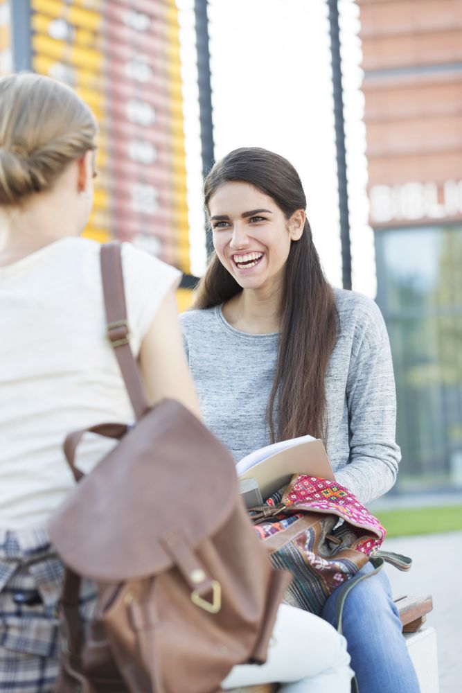 Smiling teenage girl sitting with friend on bench at university campus