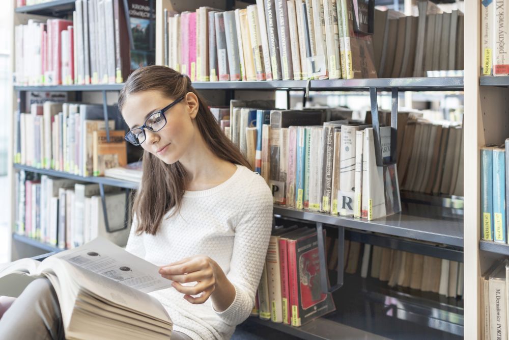 Woman reading book while sitting against bookshelf at library