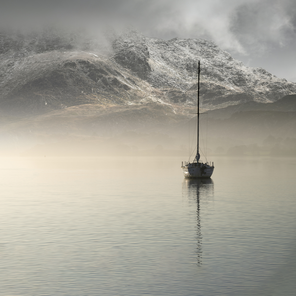 Beautiful landscape image of sailing yacht sitting still in calm lake water with mountain looming in background during Autumn Fall sunrise