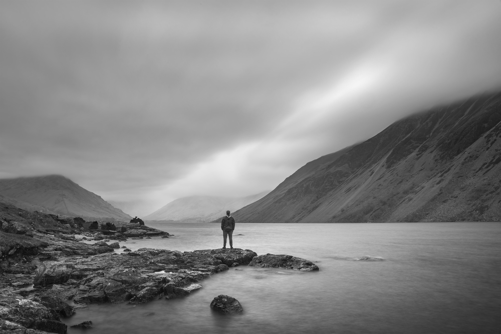 Lone figure in landscape image of Wast Water in UK Lake District during moody Spring evening in black and white