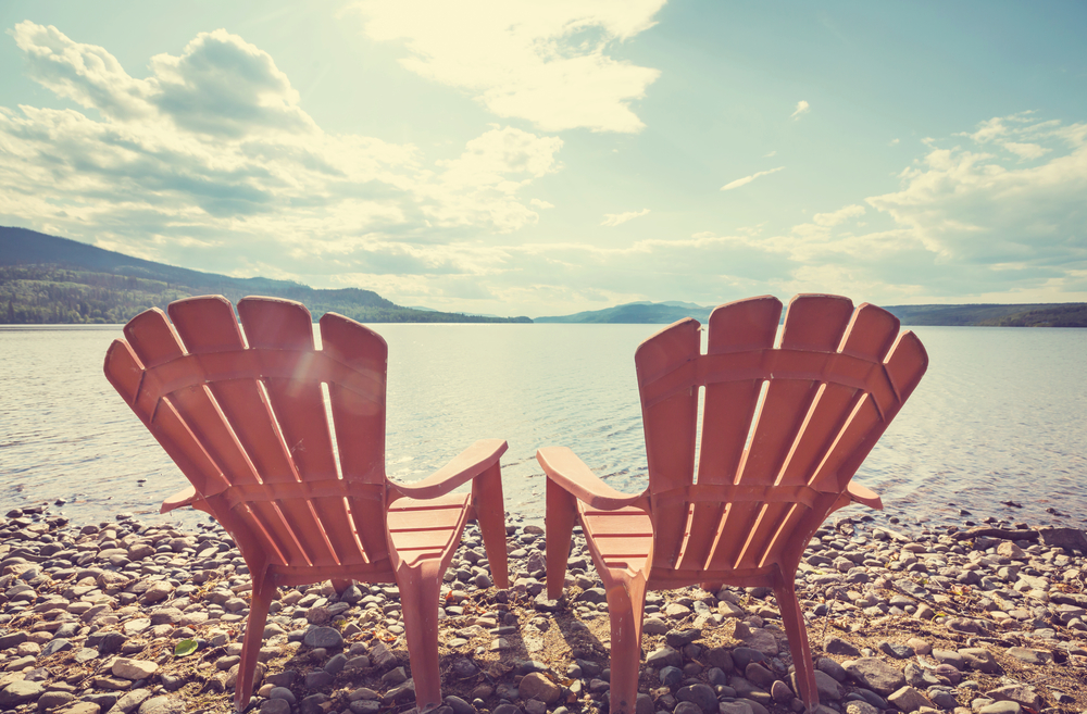 Two wooden  chairs on the shore of a lake facing the water.