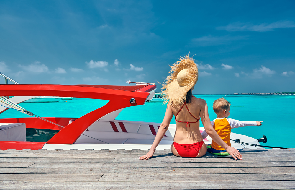Three year old toddler boy with mother sitting on wooden jetty with boat. Summer family vacation at Maldives.