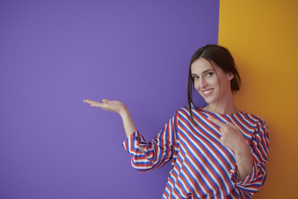 Portrait of happy smiling young beautiful woman in a presenting gesture with open palm isolated on purple and yellow background. Female model in modern fashionable clothes posing in the studio