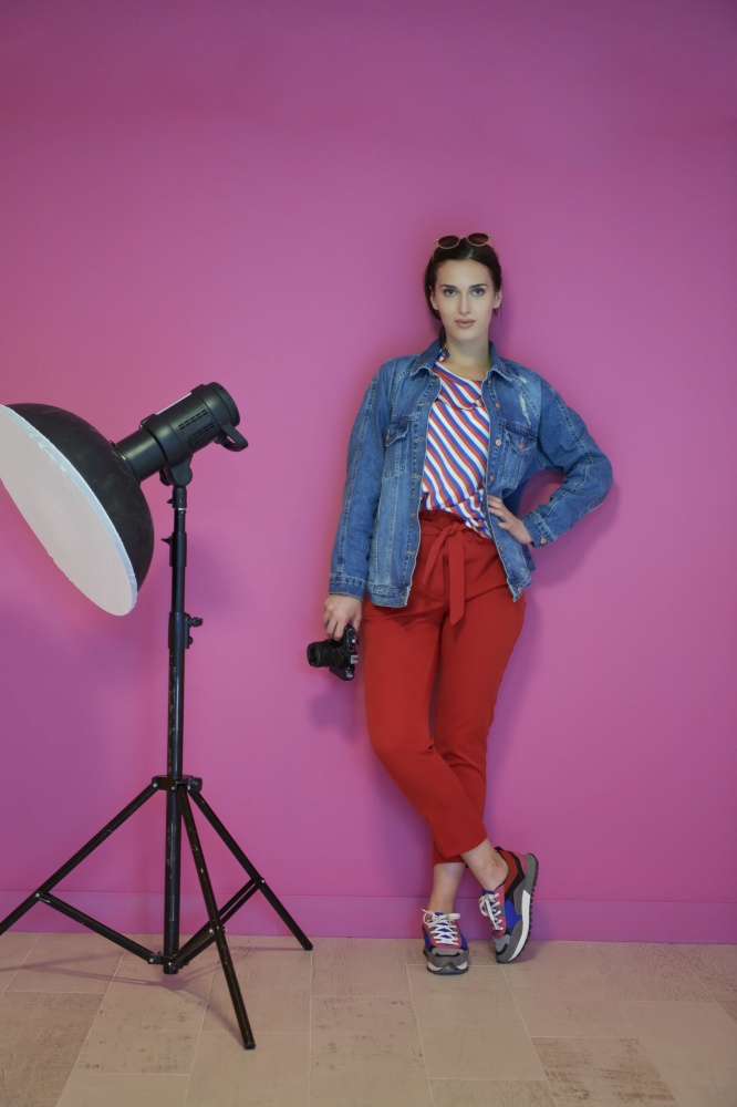 Female photographer holding camera in her hand and leaning against pink wall. Professional photography equipment in the studio