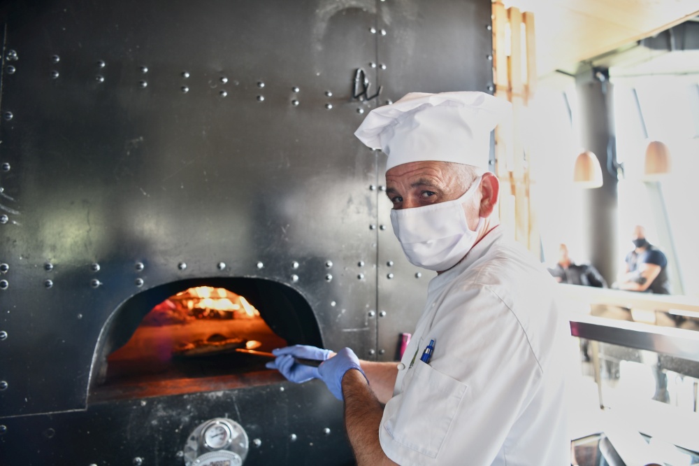Skilled chef preparing traditional italian pizza  in interior of modern restaurant kitchen with special wood-fired oven. Wearing protective medical face mask and gloves in coronavirus new normal concept