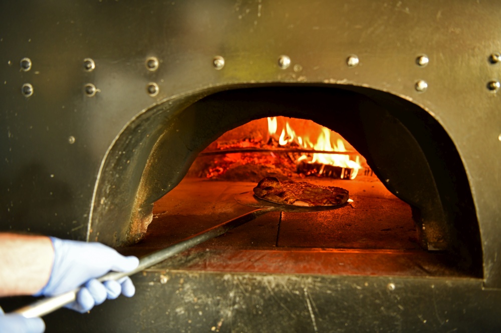 Skilled chef preparing traditional italian pizza  in interior of modern restaurant kitchen with special wood-fired oven. Wearing protective medical face mask and gloves in coronavirus new normal concept