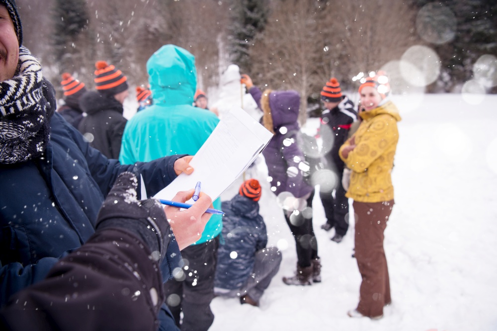 group of young happy business people after a competition measuring the height of finished snowman while enjoying snowy winter day with snowflakes around them during a team building in the mountain forest