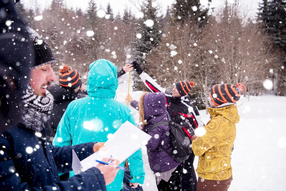 group of young happy business people after a competition measuring the height of finished snowman while enjoying snowy winter day with snowflakes around them during a team building in the mountain forest