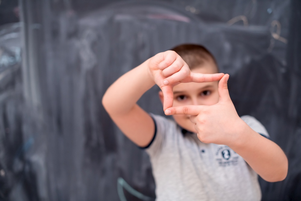 happy cute little boy having fun making hand frame gesture while standing in front of black chalkboard