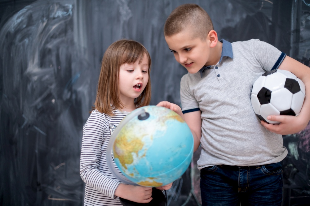 happy childrens boy with soccer ball and little girl learning about the world using globe of earth while standing in front of black chalkboard