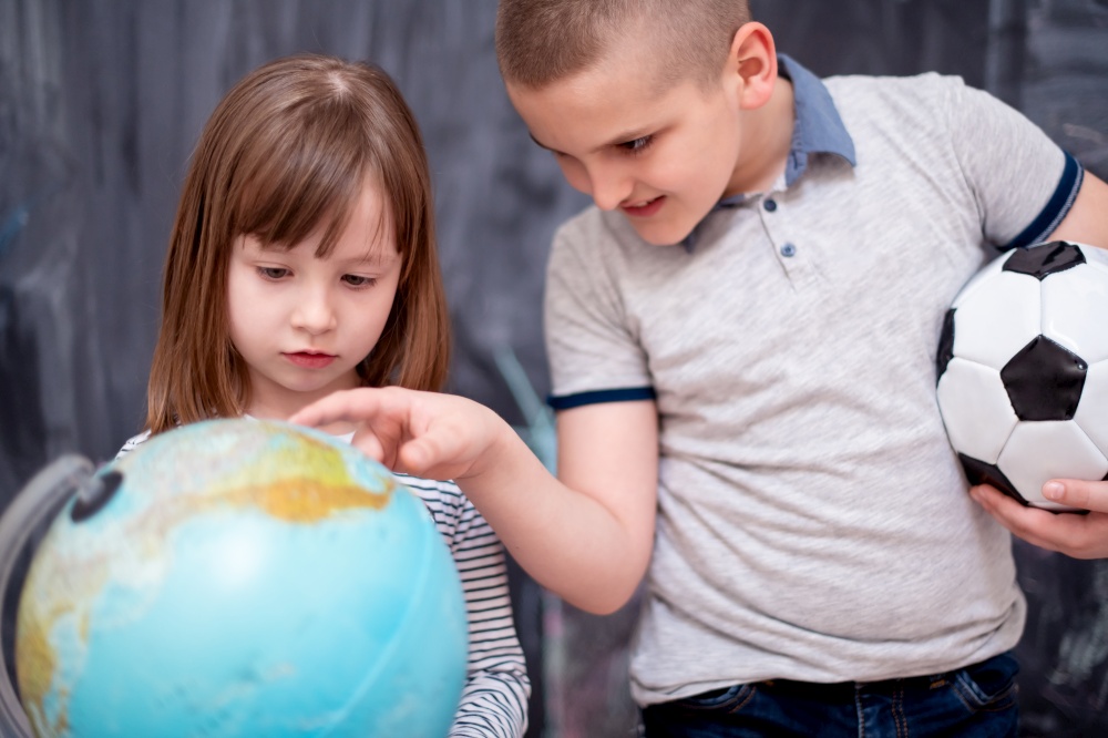 happy childrens boy with soccer ball and little girl learning about the world using globe of earth while standing in front of black chalkboard