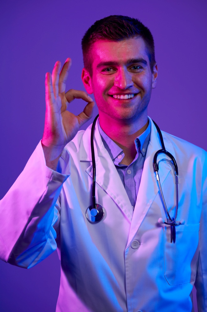 Doctor doing ok sign with fingers, excellent symbol neon lights blue and pink background. Coronavirus pandemic