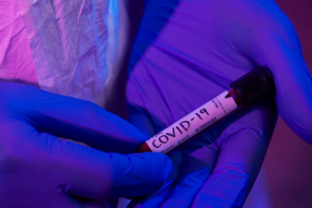 Coronavirus, Doctor holding positive covid-19 virus Blood Sample test tube. Wearing biohazard epidemic Protective mask, suit and glows neon light background