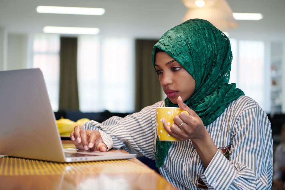 african muslim business woman wearing a green hijab drinking tea while working on laptop computer in relaxation area at modern open plan startup office. Diversity, multiracial concept