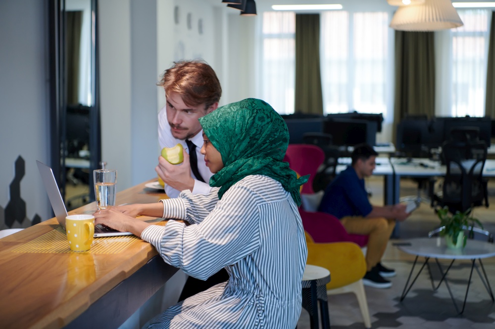 international multicultural business team.man eating apple african muslim woman wearing green hijab drinking tea while working together on laptop computer in relaxation area at modern open plan startup office