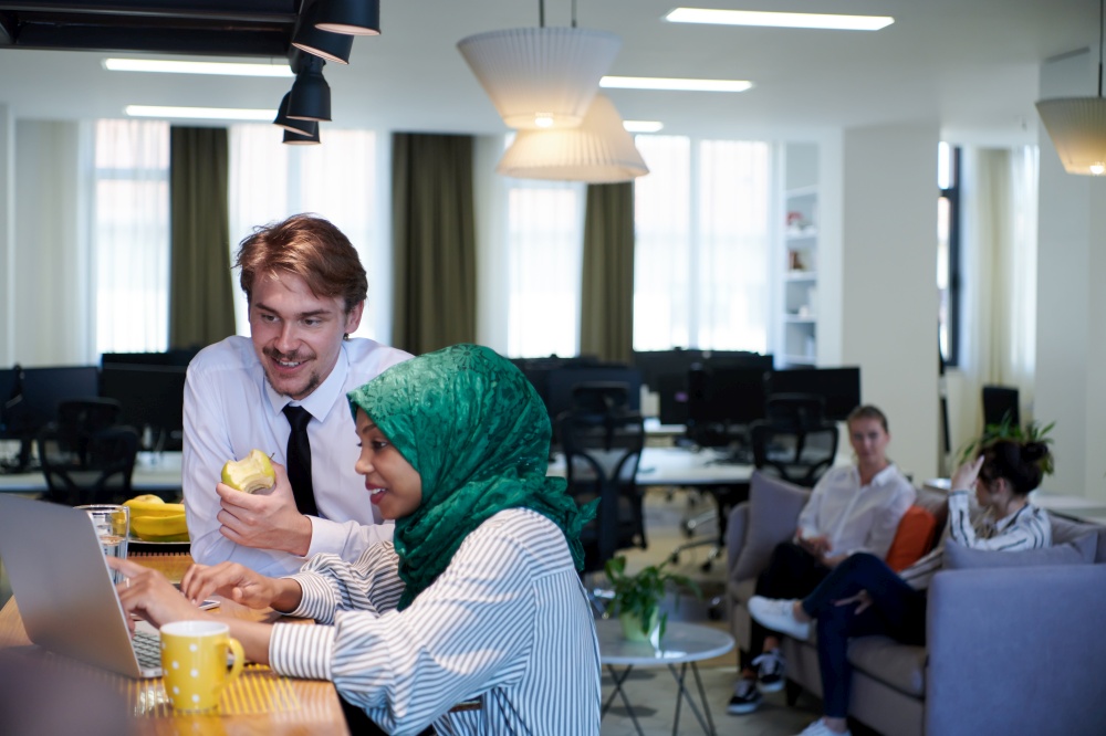 international multicultural business team.man eating apple african muslim woman wearing green hijab drinking tea while working together on laptop computer in relaxation area at modern open plan startup office