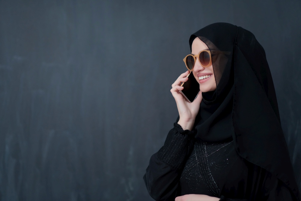 Young muslim businesswoman in traditional clothes or abaya using smartphone. Arab woman with glasses happy while standing in front of black chalkboard and representing techology, islamic  fashion and Ramadan kareem concept