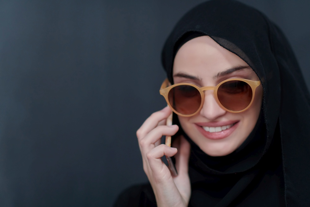 Young muslim businesswoman in traditional clothes or abaya using smartphone. Arab woman with glasses happy while standing in front of black chalkboard and representing techology, islamic  fashion and Ramadan kareem concept