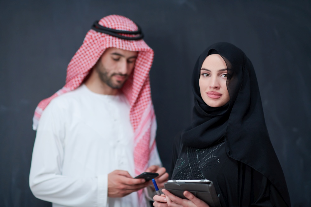 young muslim business couple arabian man with woman in fashionable hijab dress using mobile phone and tablet computer in front of black chalkboard representing modern islam fashion technology and ramadan kareem concept
