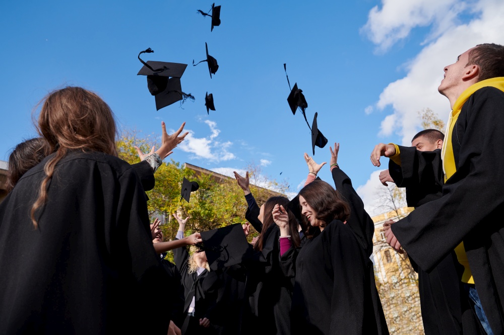 Congratulations! Low angle view of happy group of six young cheerful graduates in black gowns, throwing up their head wear in the air and celebrating, in blue summer sky, laughing, enjoying
