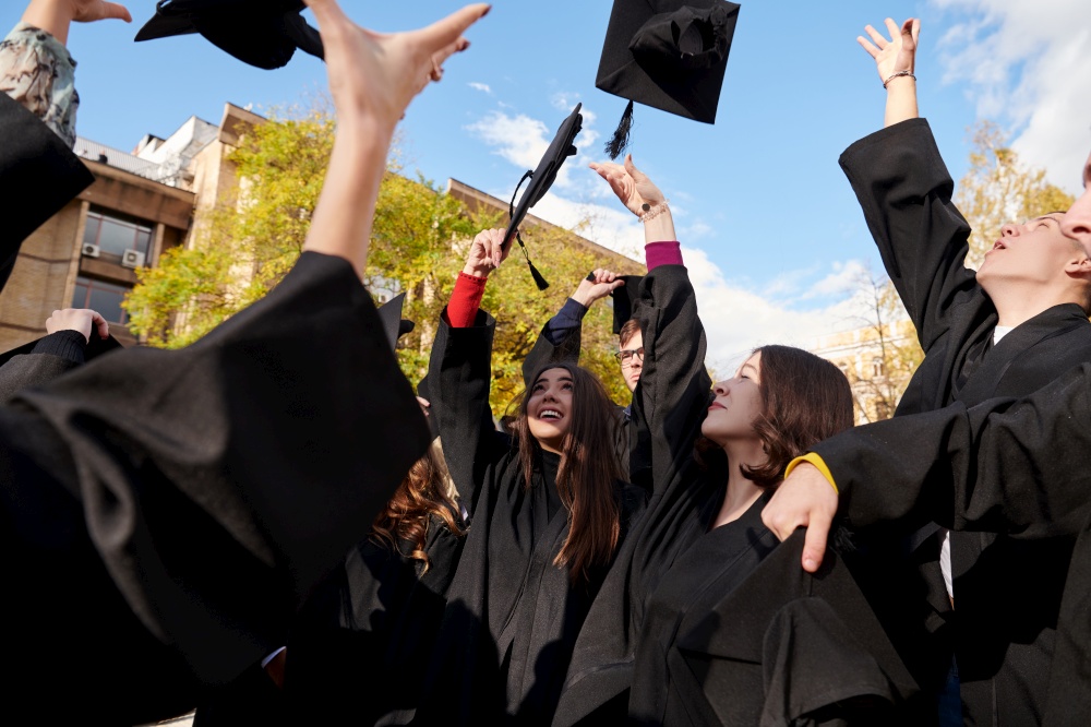 Congratulations! Low angle view of happy group of six young cheerful graduates in black gowns, throwing up their head wear in the air and celebrating, in blue summer sky, laughing, enjoying