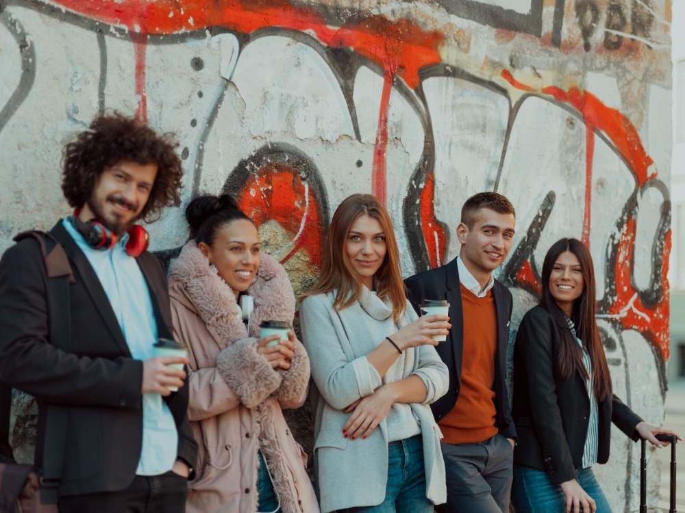 a group of diverse young businessmen on a break from work walk around the city, use smartphones, listen to music, and have fun. young people stand in front of a drawn wall in an abandoned street.