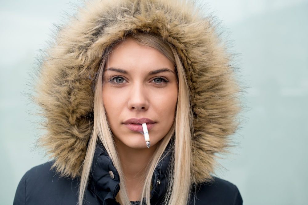portrait of young blonde girl wearing winter jacket with a hood and cigarette in the mouth isolated over white background