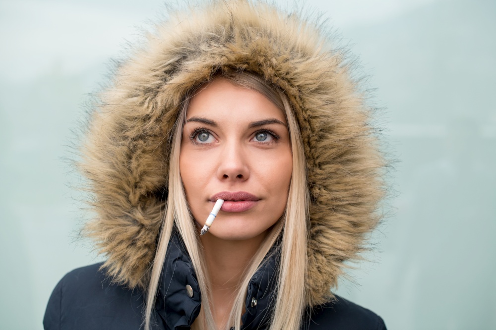 portrait of young blonde girl wearing winter jacket with a hood and cigarette in the mouth isolated over white background
