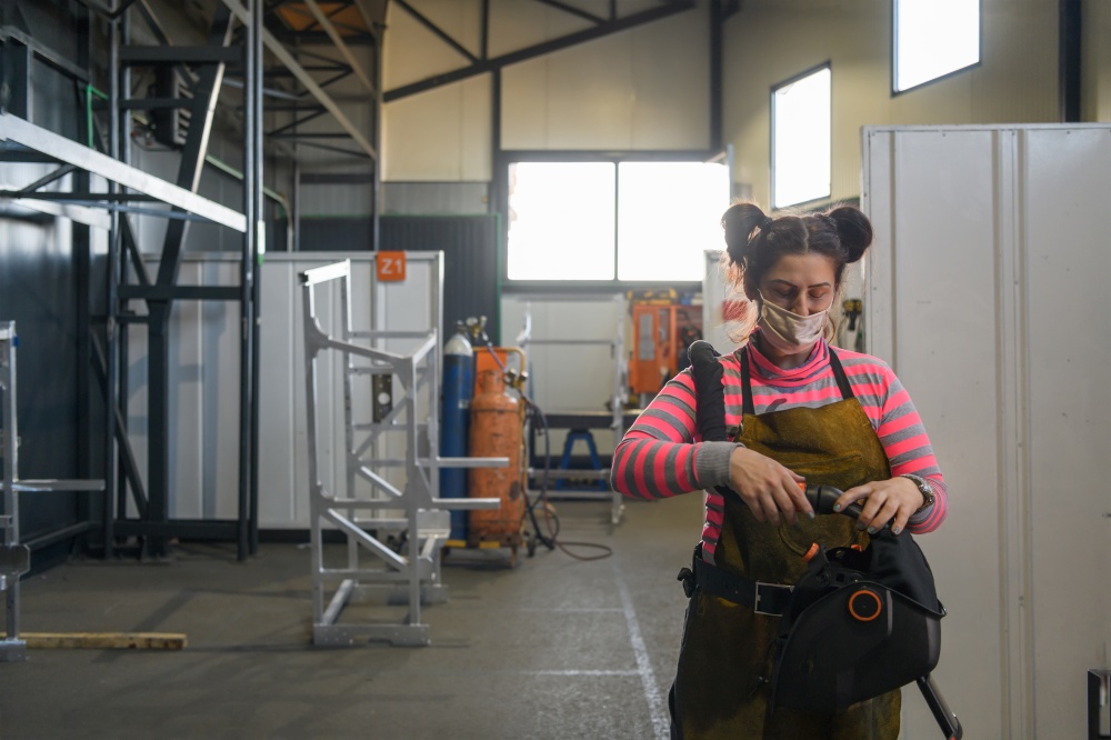A female welder preparing for a working day in the metal processing industry wears a mask on her face due to the coronavirus pandemic. High quality photo. A female welder preparing for a working day in the metal processing industry wears a mask on her face due to the coronavirus pandemic