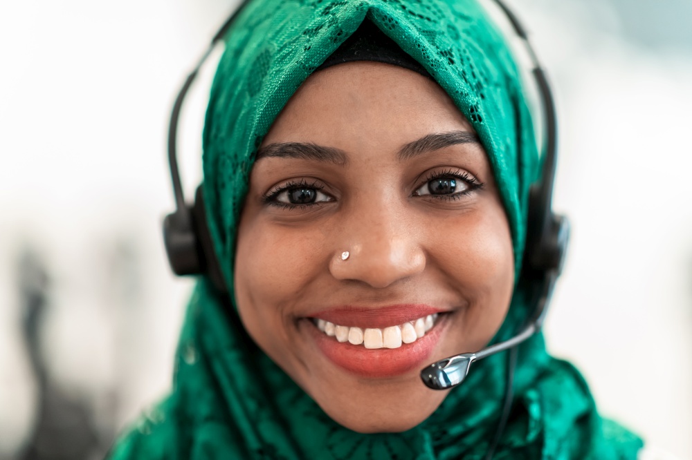 Afro Muslim female with green hijab scarf customer representative businesswoman with phone headset helping and supporting online with the customer in a modern call center. High-quality photo. Afro Muslim female with green hijab scarf customer representative business woman with phone headset helping and supporting online with customer in modern call centre