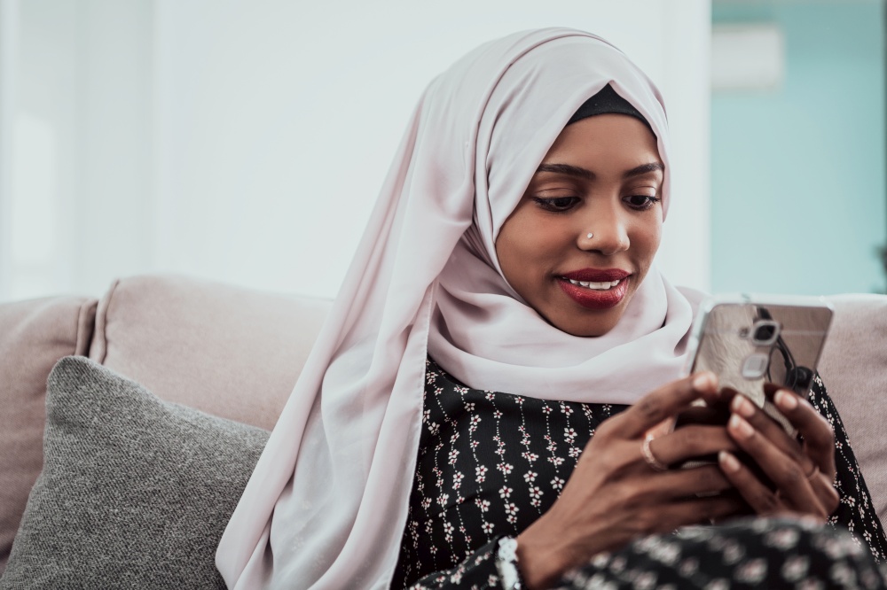 African woman using smartphone while sitting on the sofa at home wearing hijab clothes. High-quality photo. African woman using smartphone while sitting on the sofa at home wearing hijab clothes.
