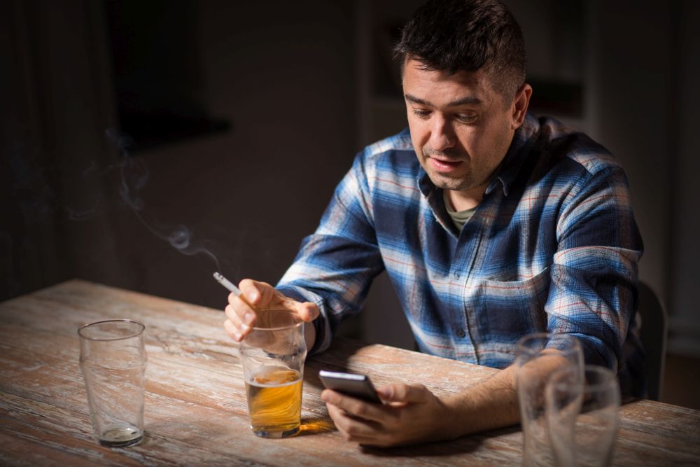 alcoholism, alcohol addiction and people concept - male alcoholic with smartphone drinking beer and smoking cigarette at night. man with cellphone drinking alcohol and smoking