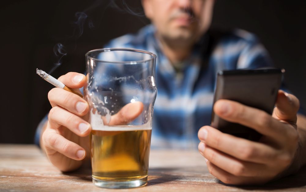 alcoholism, alcohol addiction and people concept - male alcoholic with smartphone drinking beer and smoking cigarette at night. man with cellphone drinking alcohol and smoking
