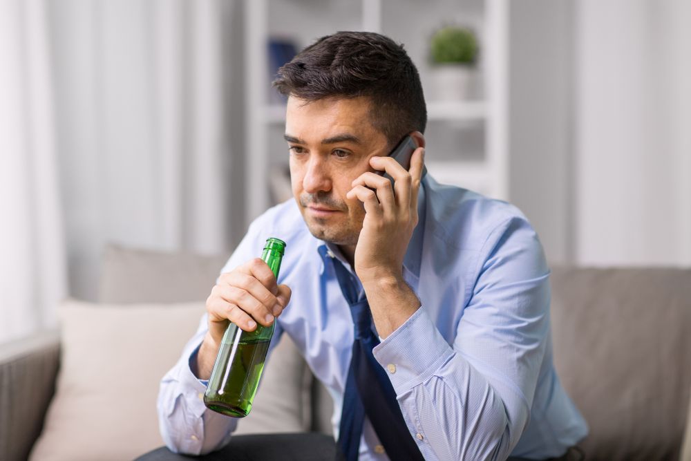 alcoholism, alcohol addiction and people concept - male alcoholic drinking beer and calling on smartphone at home. man drinking alcohol and calling on smartphone