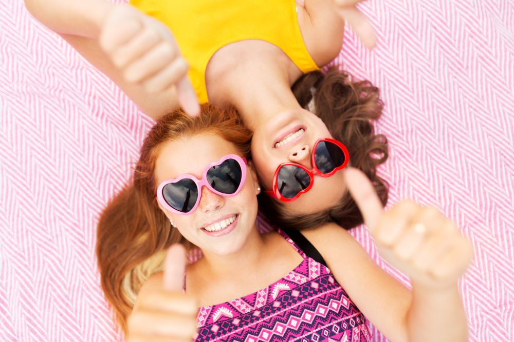 summer fashion, leisure and valentines day concept - smiling teenage girls in heart shaped sunglasses lying on picnic blanket and showing thumbs up. teenage girls in sunglasses showing thumbs up