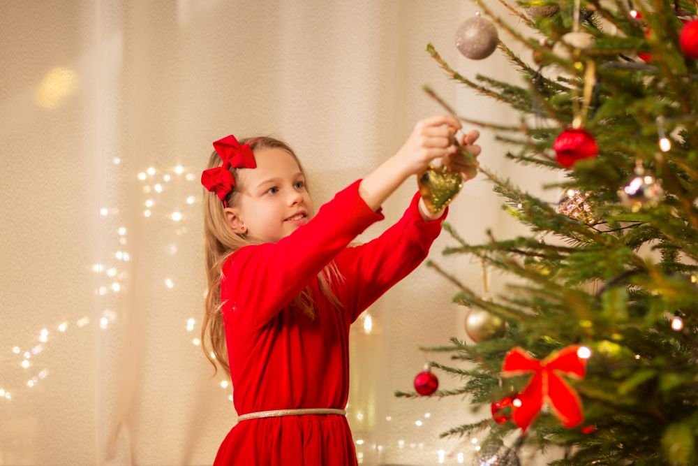 christmas, holidays and childhood concept - happy girl in red dress decorating natural fir tree. happy girl in red dress decorating christmas tree