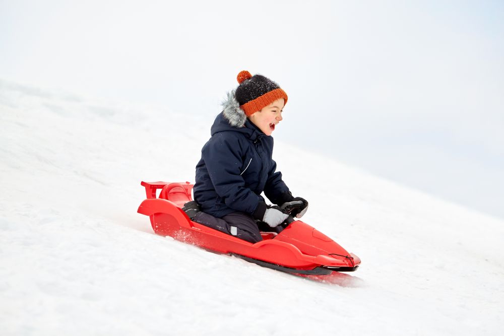childhood, sledging and season concept - happy little boy sliding on sled down snow hill outdoors in winter. happy boy sliding on sled down snow hill in winter