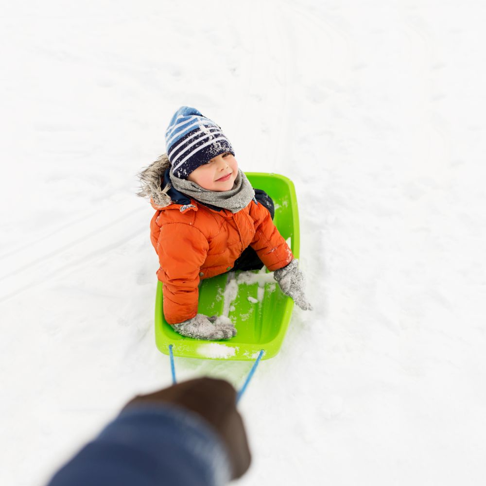 childhood, winter leisure and season concept - parent hand carrying sled with happy little boy on snow outdoors. happy boy riding sled on snow in winter