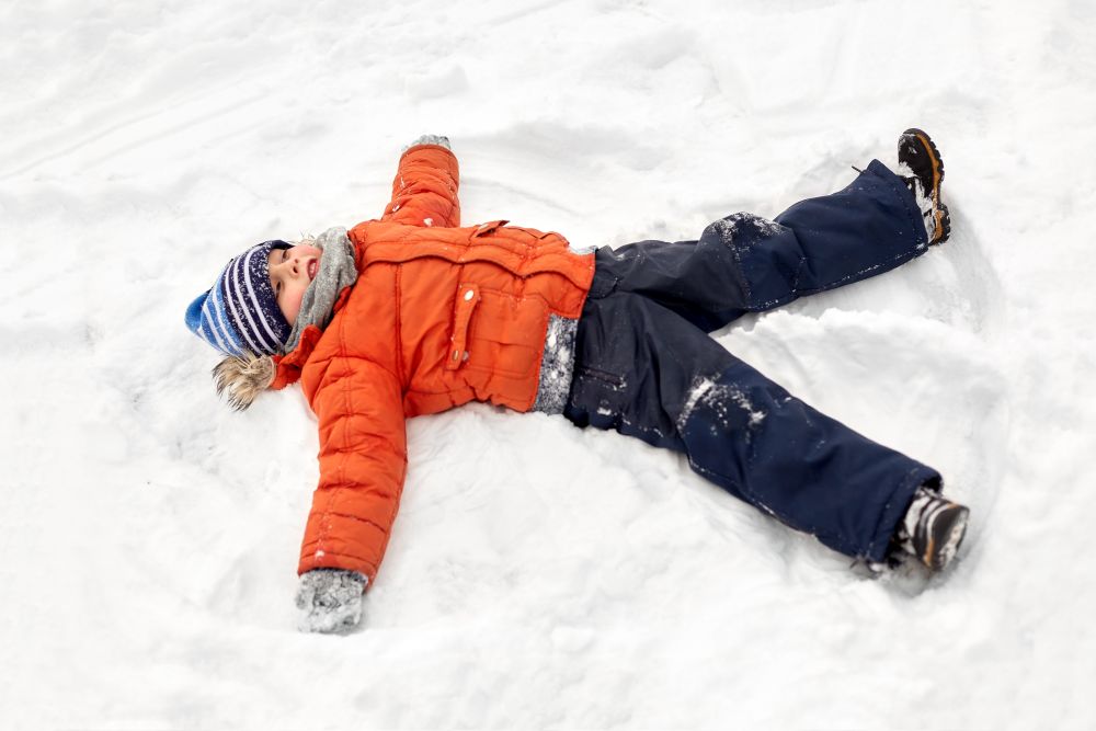 childhood, leisure and season concept - happy little boy in winter clothes making snow angels outdoors. happy little boy making snow angels in winter