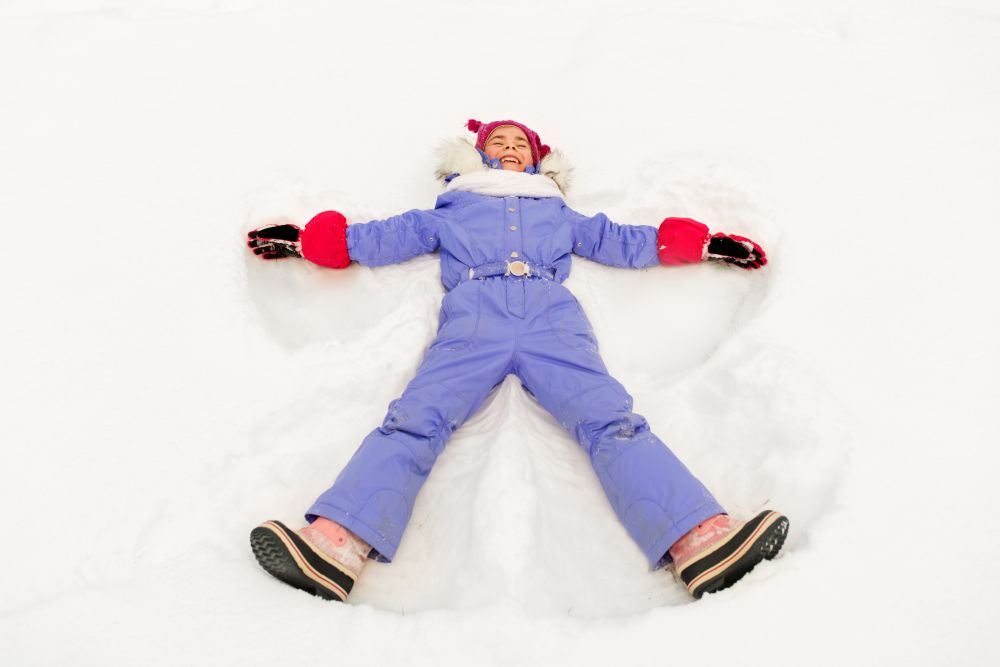 childhood, leisure and season concept - happy little girl in winter clothes making snow angels outdoors. happy little girl making snow angels in winter