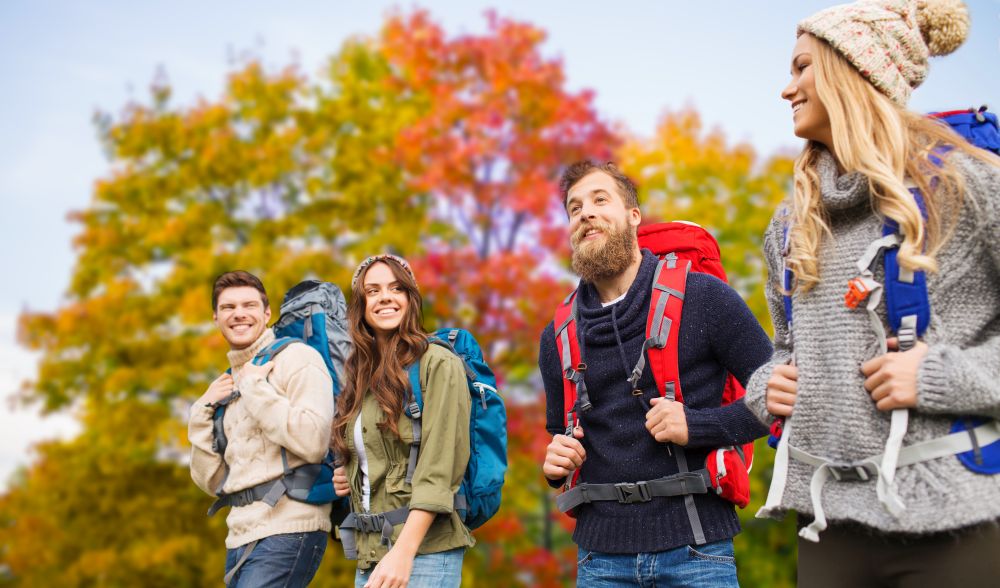travel, tourism, hike and people concept - group of smiling friends with backpacks hiking over autumn trees background. group of friends with backpacks hiking in autumn