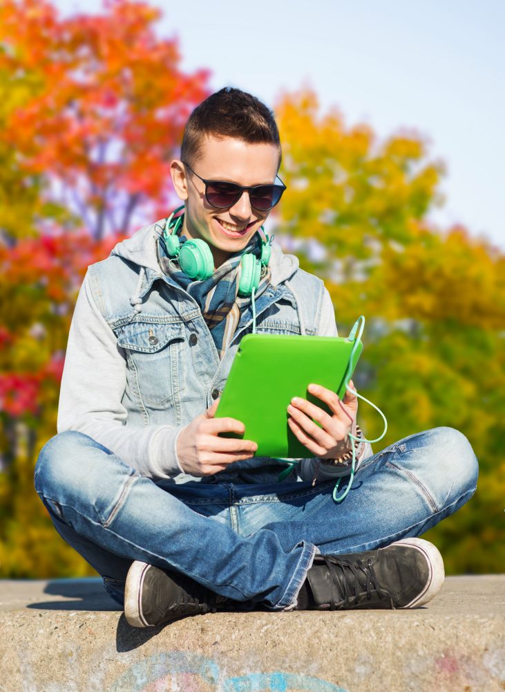 technology, lifestyle, music and people concept - smiling young man or teenage boy with tablet pc computer and headphones over autumn park background. teenage boy with tablet pc and headphones