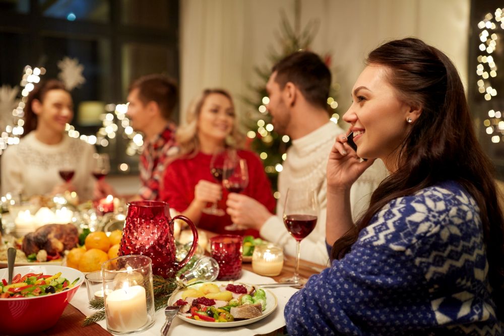 holidays, communication and celebration concept - happy young woman calling on smartphone and having christmas dinner with friends at home. woman calling on smartphone at christmas dinner