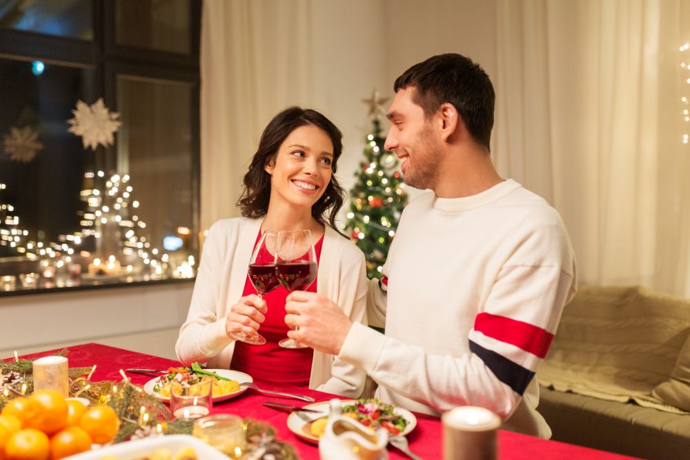 holidays, family and celebration concept - happy couple having christmas dinner at home and drinking red wine and clinking glasses. happy couple drinking red wine at christmas dinner