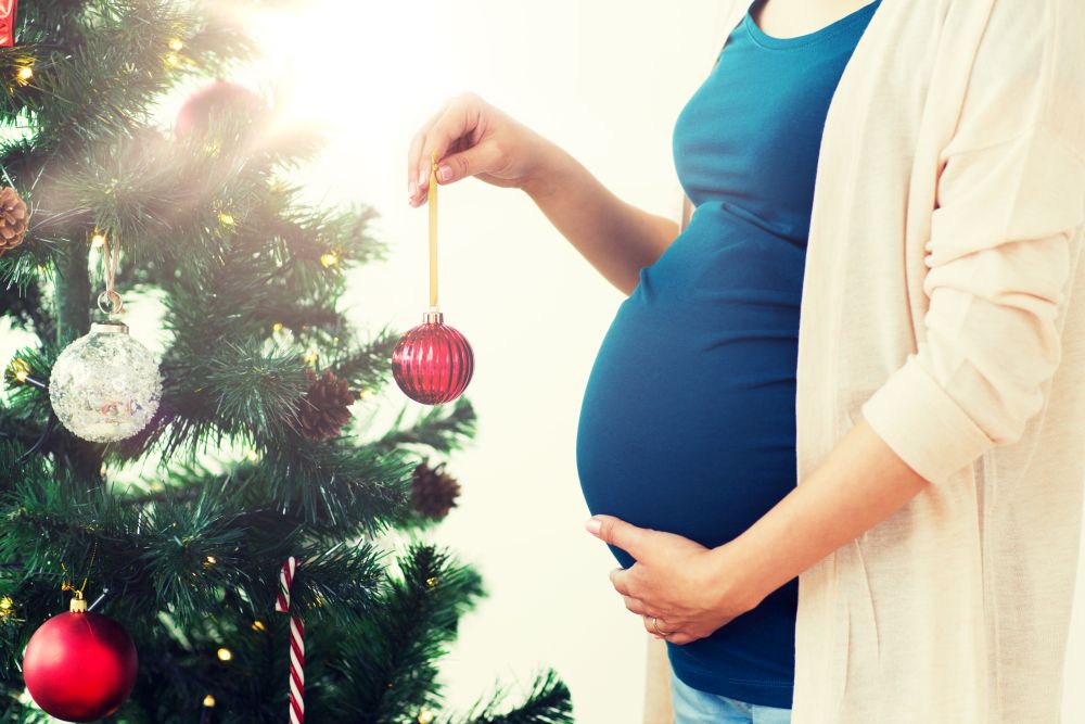 pregnancy, winter holidays and people concept - close up of pregnant woman decorating christmas tree at home. pregnant woman decorating christmas tree